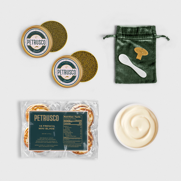 Petrusco caviar gift set with two open tins of sturgeon caviar, a pack of 16 French mini blinis, a bowl of crème fraîche, and a green velvet pouch with mother-of-pearl spoon and gold caviar key.