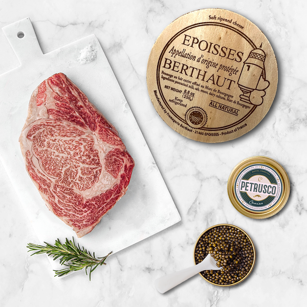 Flat lay of A5 Wagyu ribeye steak on a marble cutting board with rosemary, Berthaut Époisses AOP cheese, and an open Petrusco caviar tin with mother-of-pearl spoon.