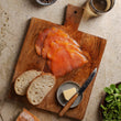 Load image into Gallery viewer, Overhead view of thinly sliced smoked salmon on a rustic wooden board with sourdough bread, butter, peppercorns, and watercress.
