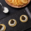 Load image into Gallery viewer, Three blini topped with crème fraîche and dark caviar on a black slate plate, with a wooden plate of blini and a spoon of crème fraîche in the background.
