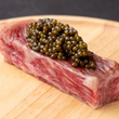 Load image into Gallery viewer, A thick cut of marbled A5 Wagyu beef topped with a generous mound of dark olive-green caviar on a wooden board.
