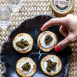 Load image into Gallery viewer, Caviar on crackers with a hand pointing at one of them, on a dark stone platter with glasses and a jar in the background.
