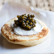 Load image into Gallery viewer, Blini topped with crème fraîche and a spoonful of dark Amur caviar pearls, close-up on a white plate.
