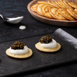 Load image into Gallery viewer, Two blini topped with crème fraîche and dark ossetra caviar on a black slate plate, with a wooden board of blini and a spoon in the background
