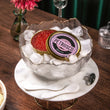 Load image into Gallery viewer, Petrusco Smoked Trout Roe tin on ice in a gold-rimmed glass bowl on a marble stand with mother-of-pearl spoons, pink flower, and green velvet backdrop.
